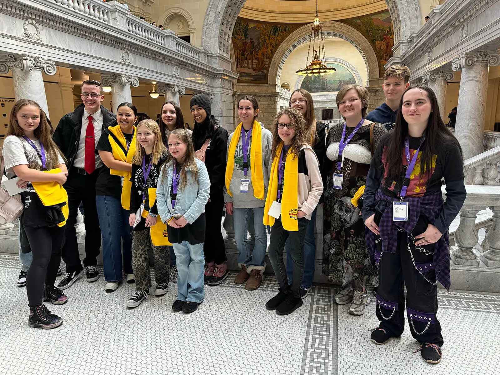 A diverse group of people posing in a grand, ornately decorated building with marble columns. They wear yellow scarves, some with lanyards, and smile, conveying a joyful and united atmosphere.